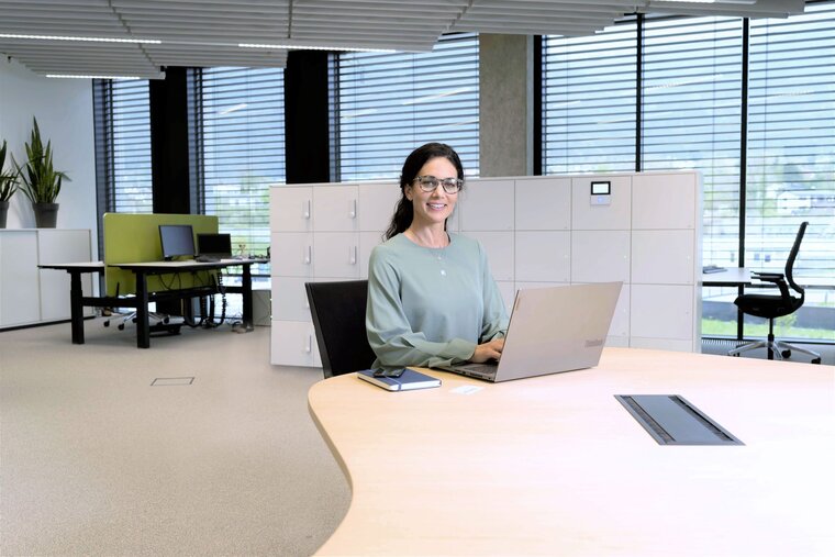 Eine Damen mit Brille sitzt an einem großen Besprechungstisch vor ihrem Laptop. Hinter ihr sind weitere Arbeitspätze du ein modernes, in Weiß gehaltenes Gantner Schließsystem zu erkennen.