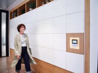 Eine Frau mit Brille und lockigem Kurzhaarschnitt geht zielgerichtet an einer langen Wand mit Schließfächern in weißem und hölzernen Dekor vorbei, an derenen Ende sich moderne und intuitive Interface von Gantner befindet. 