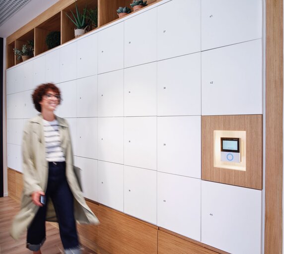 Eine Frau mit Brille und lockigem Kurzhaarschnitt geht zielgerichtet an einer langen Wand mit Schließfächern in weißem und hölzernen Dekor vorbei, an derenen Ende sich moderne und intuitive Interface von Gantner befindet. 