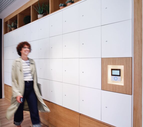 Eine Frau mit Brille und lockigem Kurzhaarschnitt geht zielgerichtet an einer langen Wand mit Schließfächern in weißem und hölzernen Dekor vorbei, an derenen Ende sich moderne und intuitive Interface von Gantner befindet. 