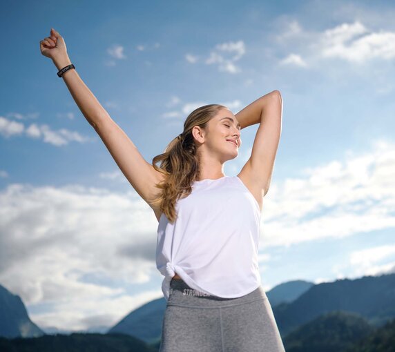 Eine sportlich gekleidete Frau steht unter freiem Himmel und streckt ihre Arme genüsslich von sich weg.