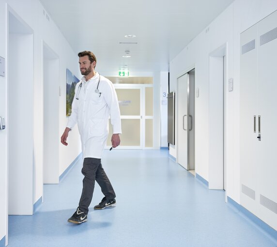 Ein Mann im Arztkittel mit einem Mitarbeiterausweis in der Hand geht auf eine Tür im Krankenhaus zu.