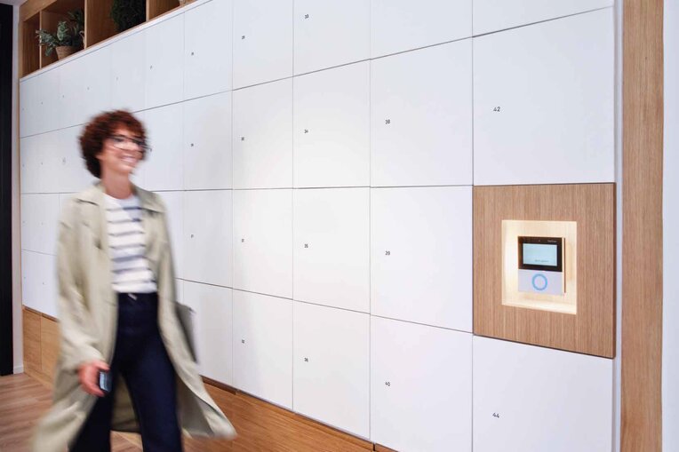 Eine Frau mit Brille und lockigem Kurzhaarschnitt schlendert zielstrebig an einer langen Wand mit Schließfächern in Weiß und Holzdekor vorbei, an deren Ende eine moderne, intuitiv zu bedienende Schnittstelle von Gantner steht. 